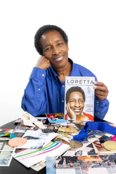 Loretta smiling with a copy of her book, Loretta, over a table of medals and photos