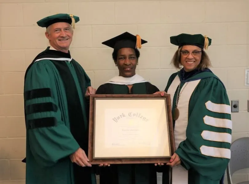 Loretta in cap and gown holding a framed York College degree between two faculty members in green robes